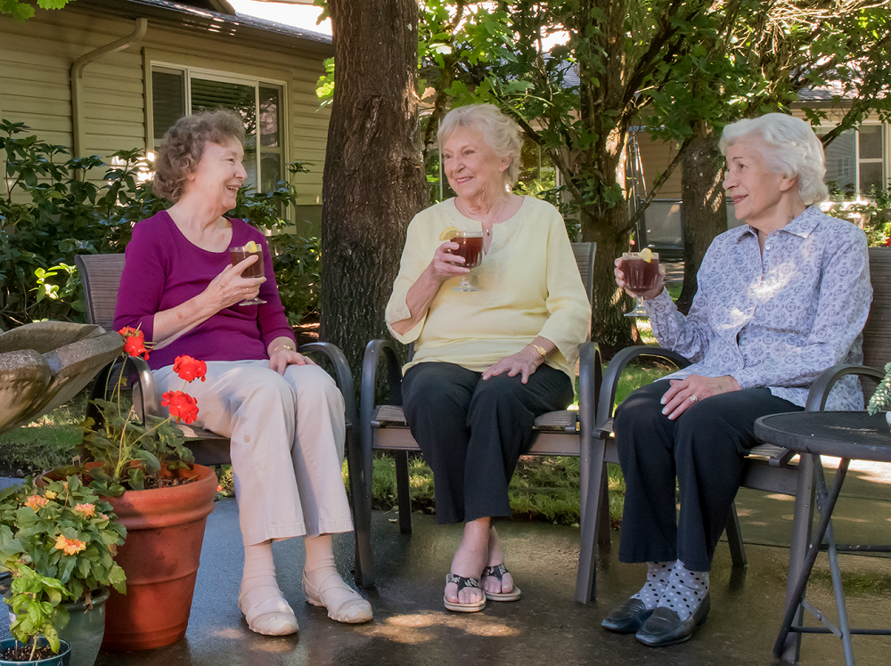 Three seniors enjoying drinks and conversation in a garden setting.
