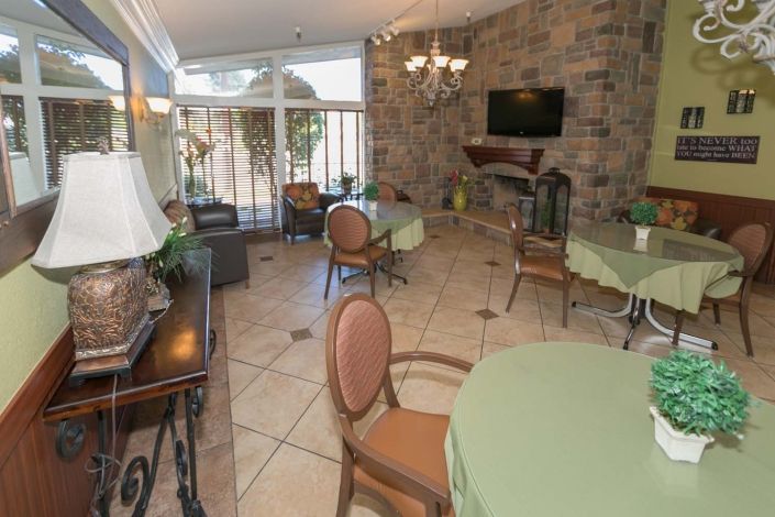 A cozy dining area with tables and chairs, featuring a stone wall, large windows, and a television.