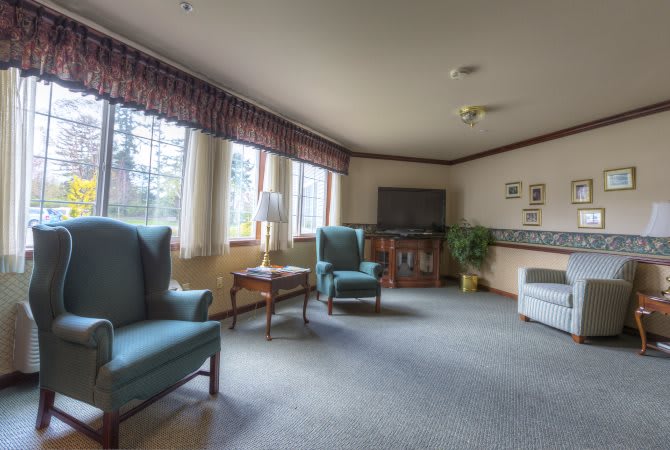 A cozy living room with large windows, two armchairs, a television, and carpeted flooring.