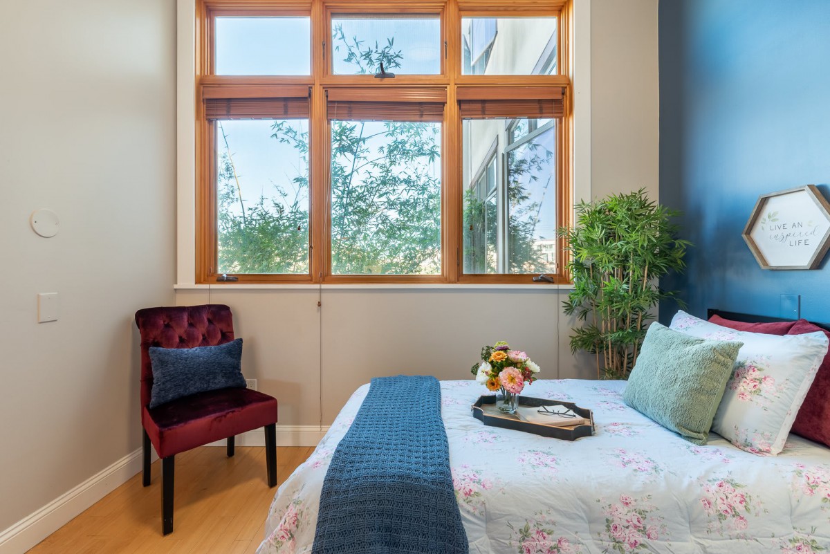 A cozy bedroom with a large window, featuring a bed adorned with floral bedding, a side chair, and vibrant decorative elements.