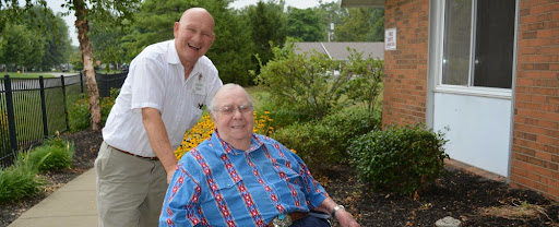 Two men enjoying a sunny day outside, with one seated and the other standing behind him.