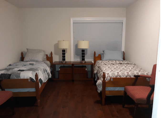 A cozy bedroom with two twin beds, each adorned with patterned bedding and separated by a nightstand with two lamps. The room features a large window and hardwood flooring.