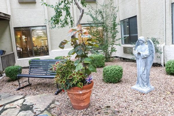 A peaceful courtyard featuring a potted plant, a statue, and a bench surrounded by gravel and small bushes.