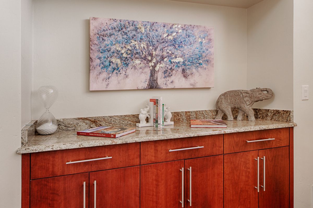 A kitchen area featuring wooden cabinets with a granite countertop, adorned with decorative items and a tree-themed artwork on the wall.