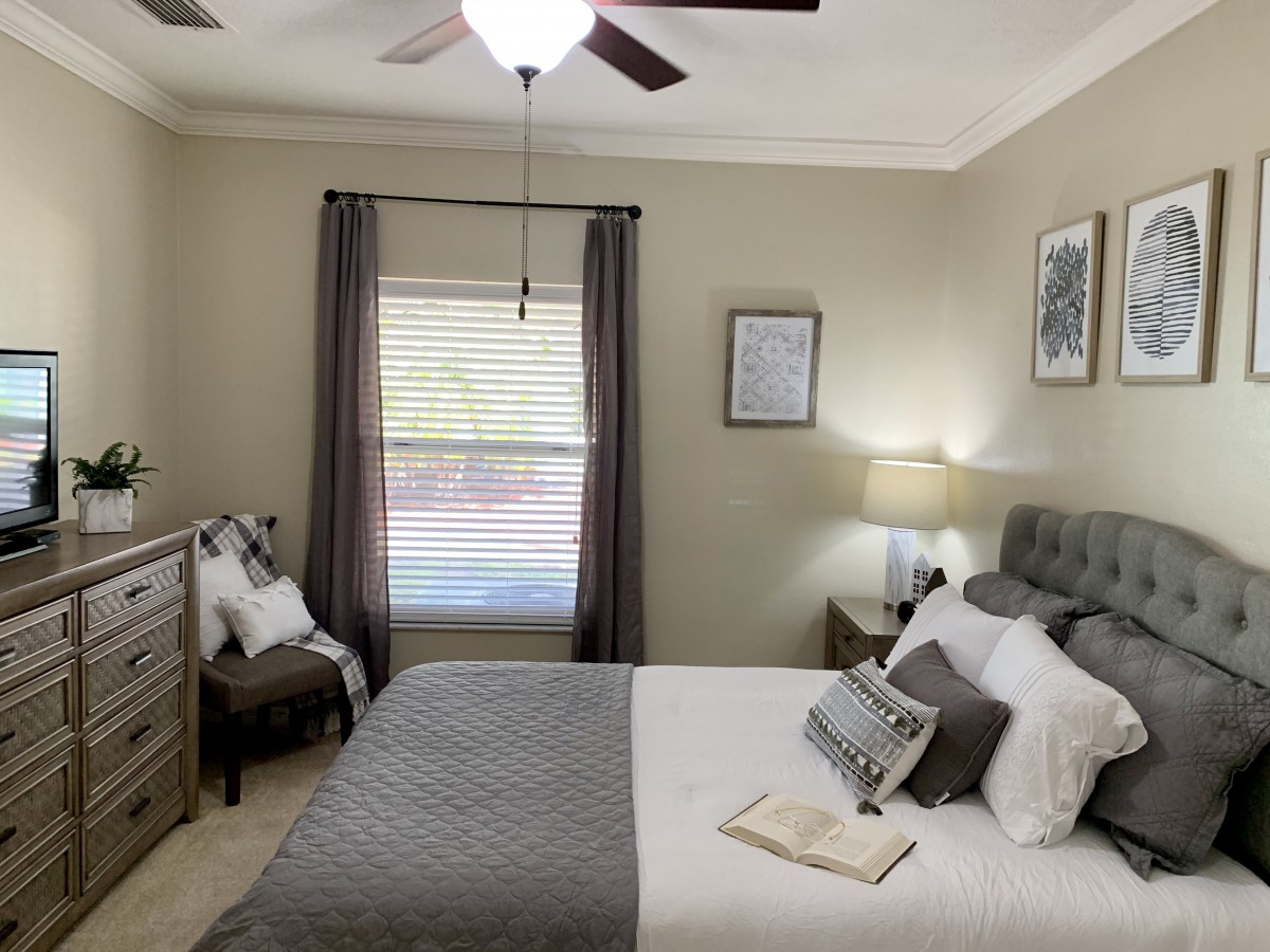 A cozy bedroom with a neutral color scheme, featuring a bed with gray bedding, a bedside lamp, and natural light filtering through the curtains.