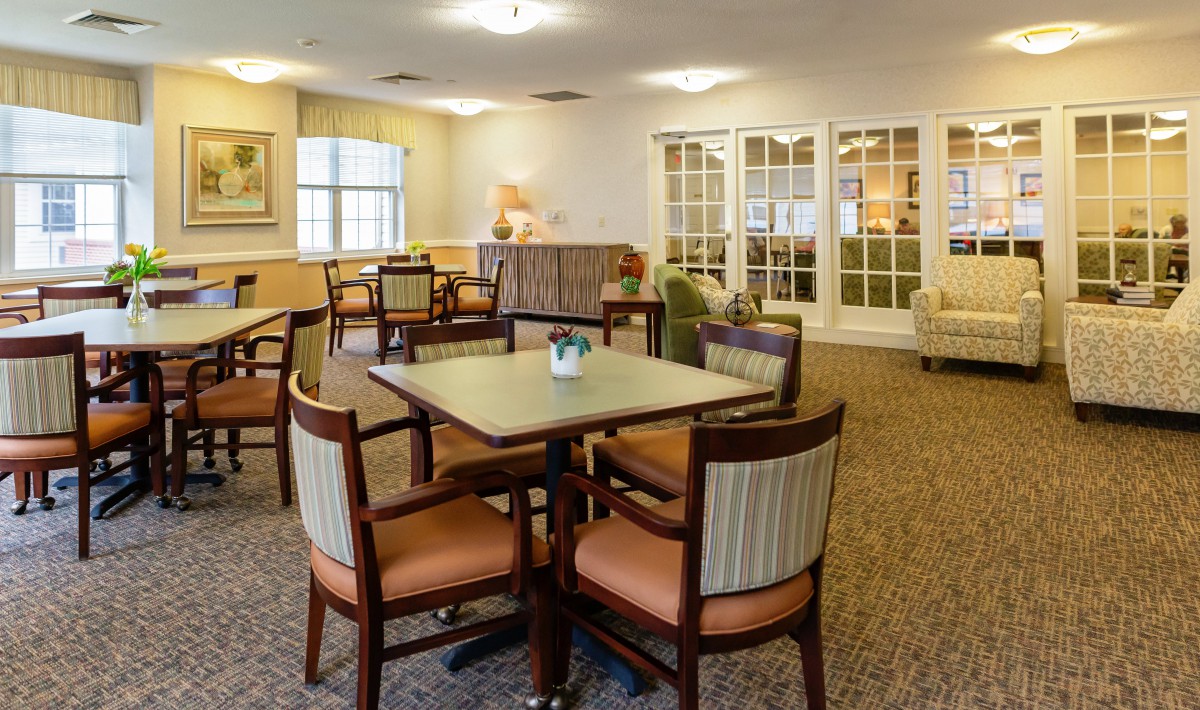 A cozy dining area with tables and chairs arranged on a carpeted floor, illuminated by overhead lighting.