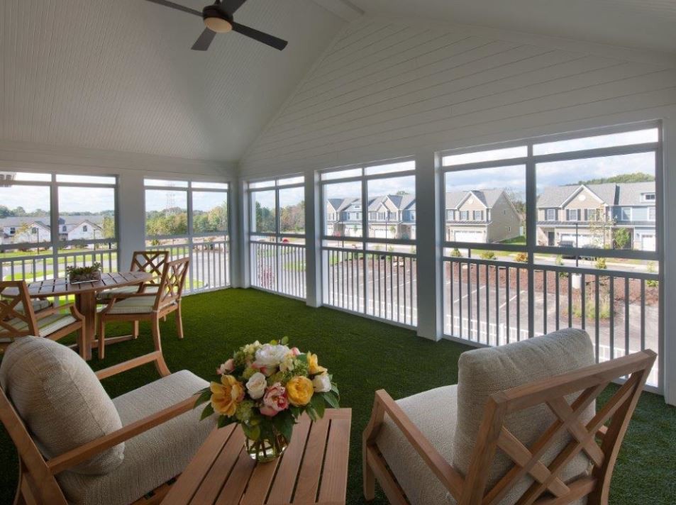 A spacious porch area with comfortable seating and a table adorned with flowers, surrounded by large windows offering a view of the neighborhood.