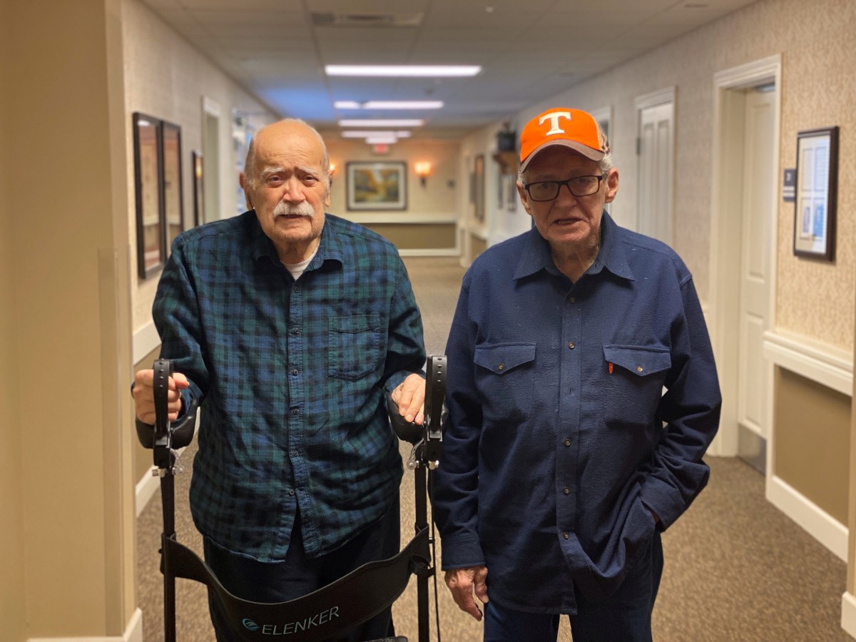 Two elderly men walking in a hallway, one using a walker.