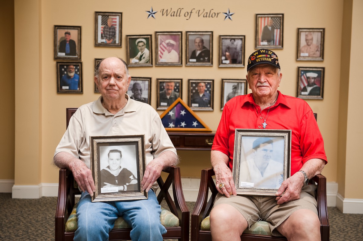 Two elderly men holding framed photographs, seated in front of a 'Wall of Valor' with various military portraits displayed.