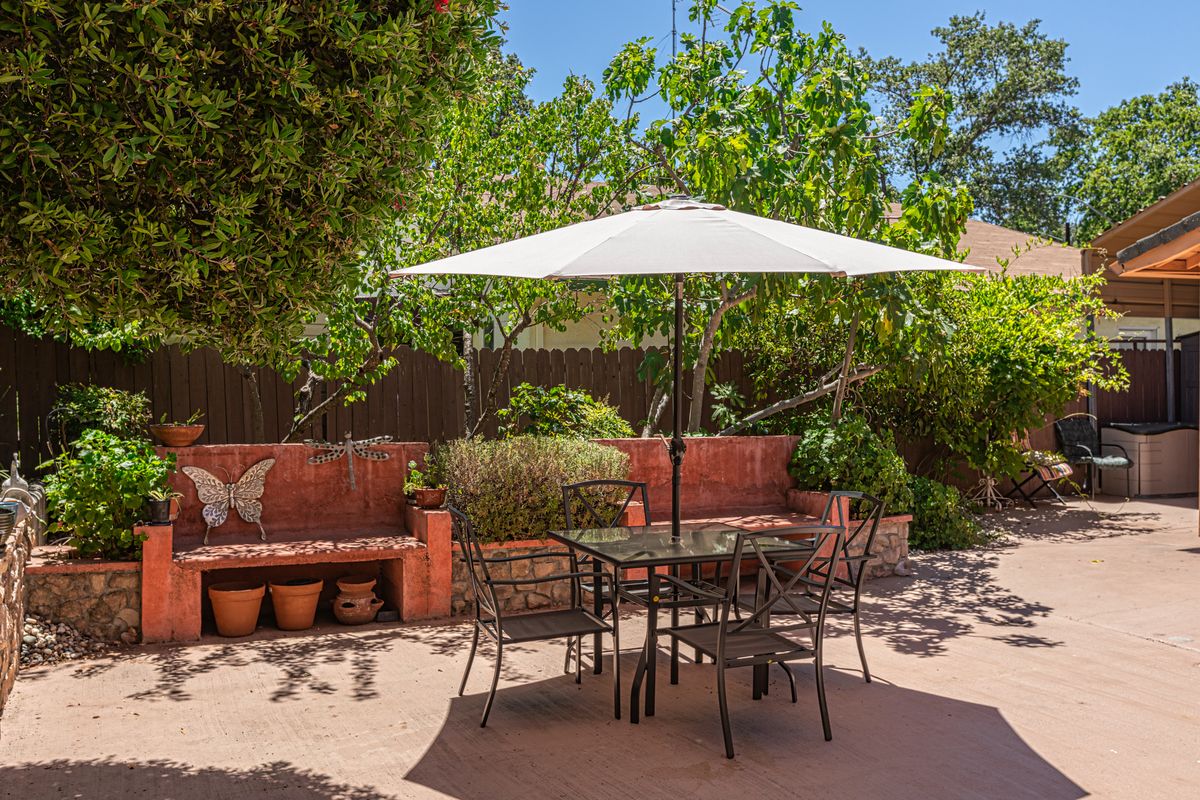 A sunny patio with a table set and umbrella, surrounded by greenery and a brick wall.