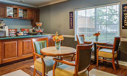 A cozy dining area with wooden furniture, decorated with vases of flowers, and illuminated by natural light from a window.