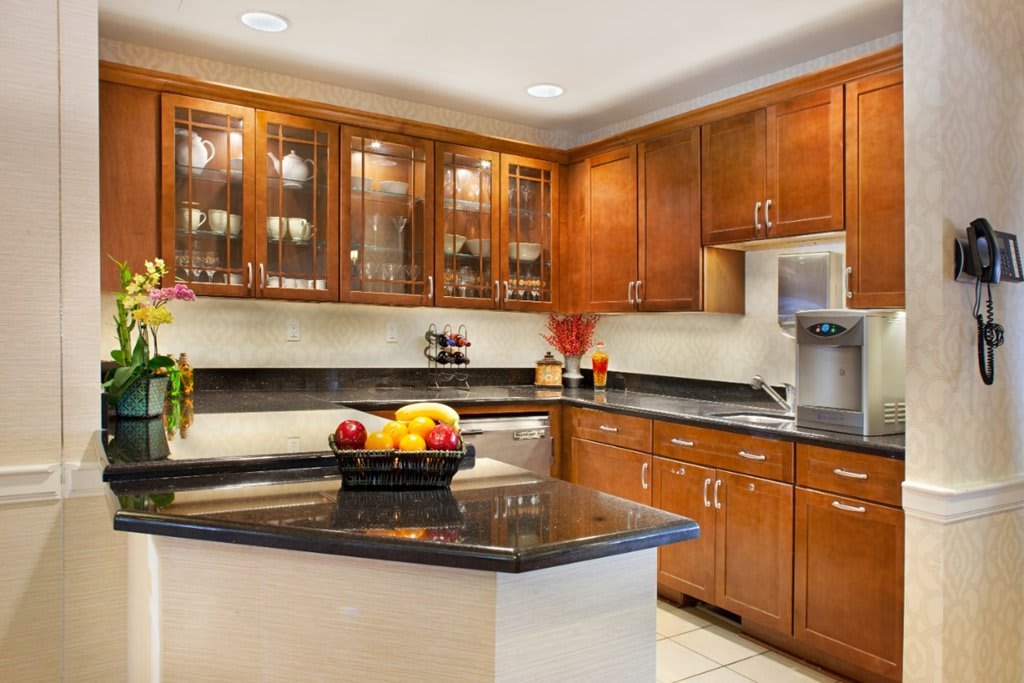 A modern kitchen with wooden cabinets and granite countertops, featuring a central island and a bowl of fruit.