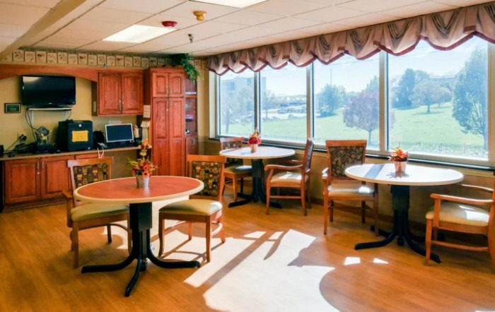 A cozy dining area with tables and chairs, bathed in natural light from large windows.
