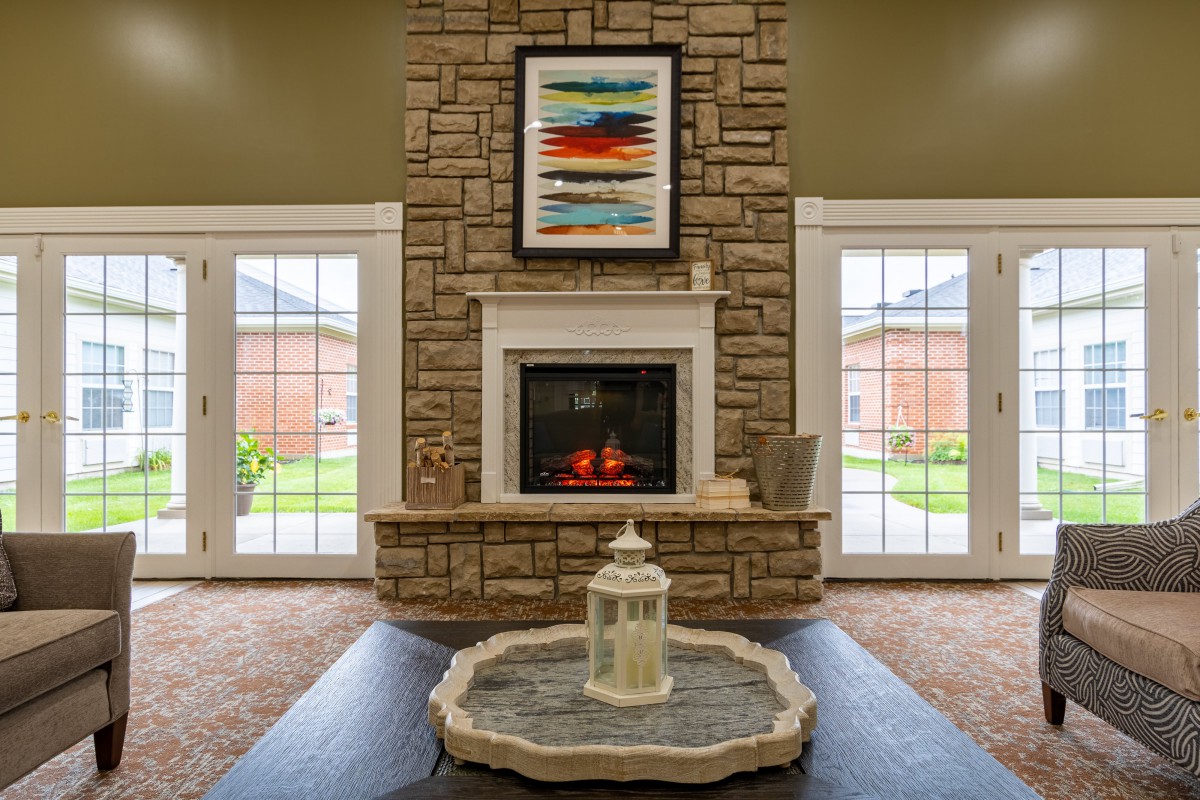 A cozy living room with a central stone fireplace, flanked by two large windows and decorated with colorful artwork.