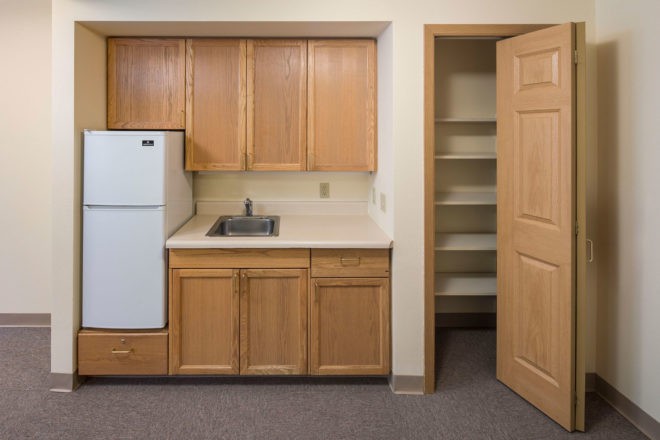 A small kitchen with wooden cabinets, a refrigerator, and an open pantry door.