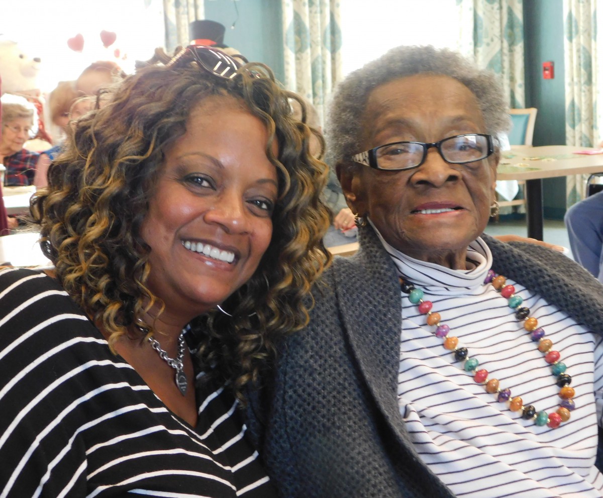 Two people smiling indoors, one wearing glasses and beads.