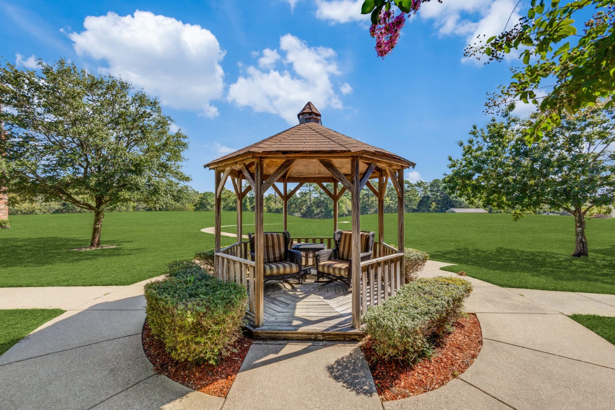 A wooden gazebo surrounded by lush greenery and a paved walkway on a sunny day.