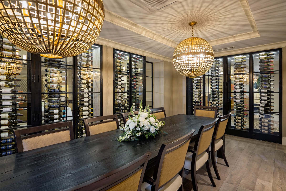 An elegant dining room with a luxurious wine cellar, chandeliers, and a long table adorned with flowers.