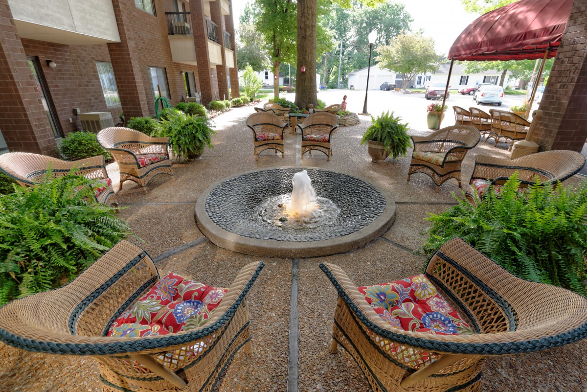 A cozy outdoor patio with wicker chairs arranged around a central fountain, surrounded by lush green plants.