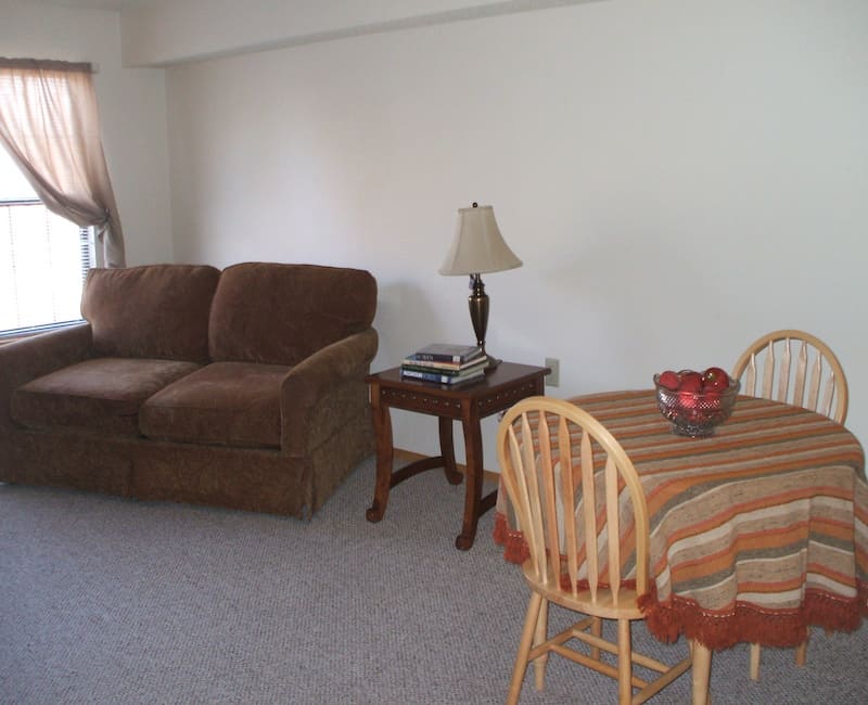 A cozy living room with a brown sofa, side table with a lamp, and a dining table with chairs.