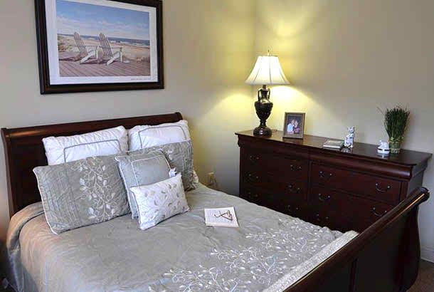 A cozy bedroom with a neatly made bed, decorative pillows, a lamp on a dresser, and framed artwork on the wall.