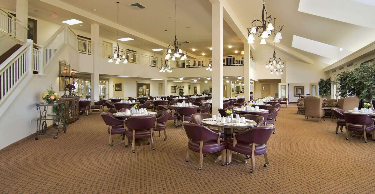A spacious dining room with multiple round tables and chairs, featuring chandeliers and a carpeted floor.