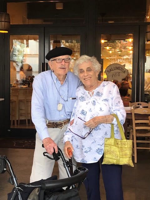 An elderly couple standing together in front of a store, one holding a cane.