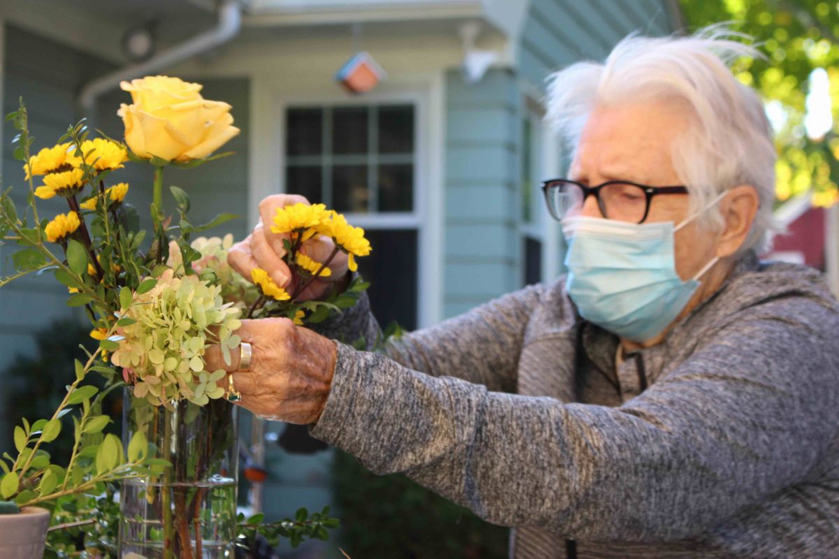 An elderly woman arranges flowers while wearing a mask outdoors.
