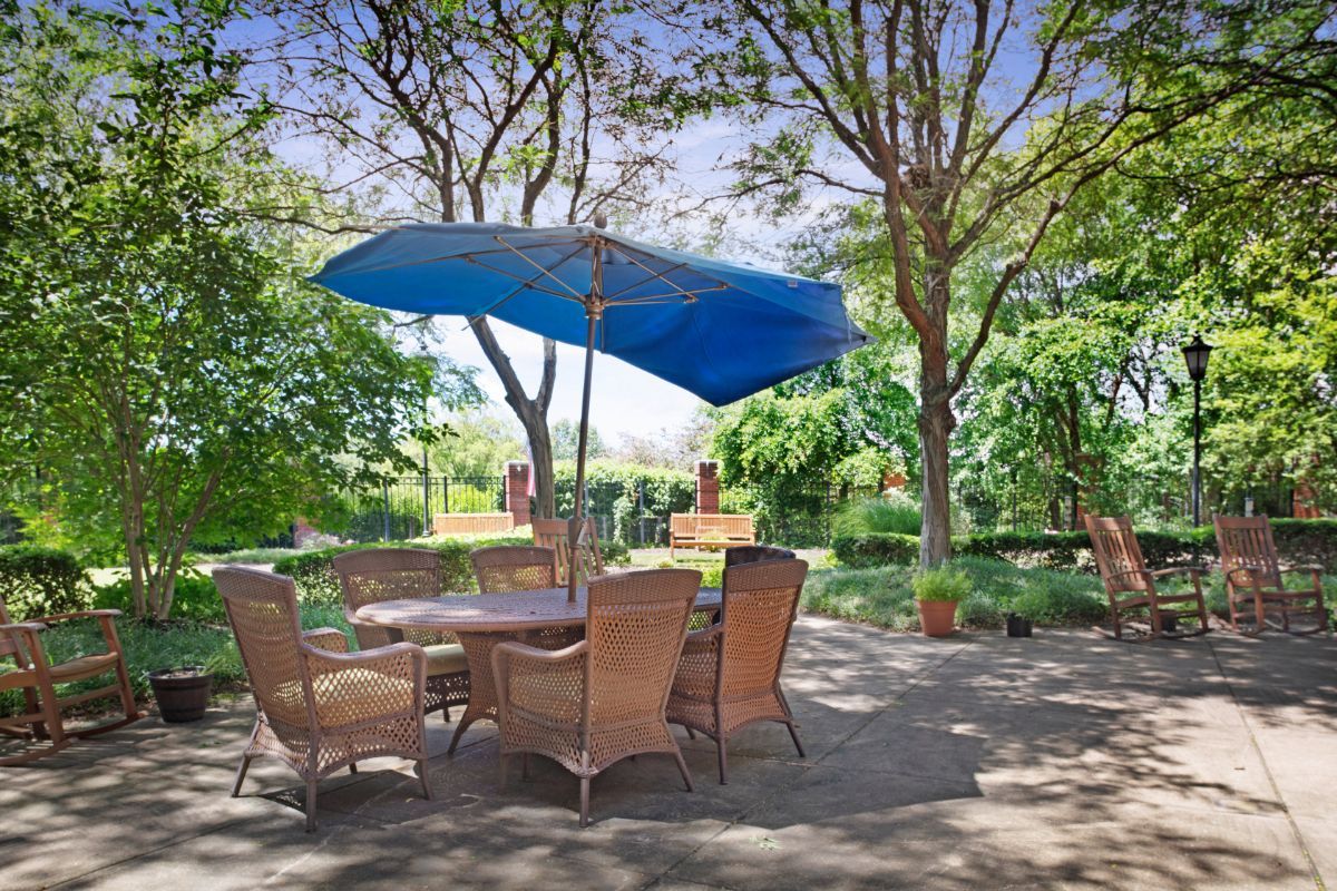 A peaceful outdoor patio with wicker chairs and a table under a blue umbrella, surrounded by lush greenery.