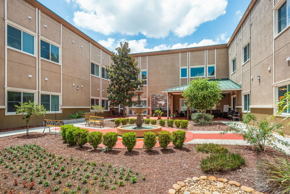 A well-maintained courtyard garden with neatly trimmed shrubs and a central pathway, located in front of a two-story building under a partly cloudy sky.