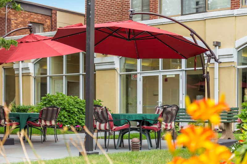 A cozy outdoor patio area with red umbrellas, chairs, and a table, surrounded by greenery and flowers.