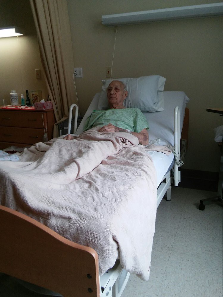 An elderly patient lies in a hospital bed, covered with a blanket.