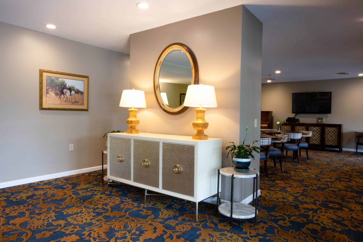 A modern living room interior with a sideboard, decorated with two lamps and an oval mirror above. The room features a patterned carpet and additional seating in the background.