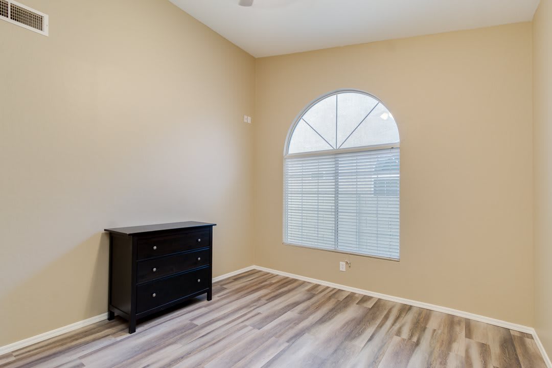An empty room with wood flooring, beige walls, a small dresser, and an arched window.