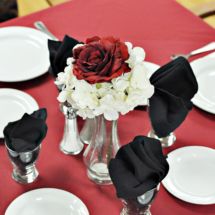 An elegant table setting featuring a red rose and white flower centerpiece with black napkins.