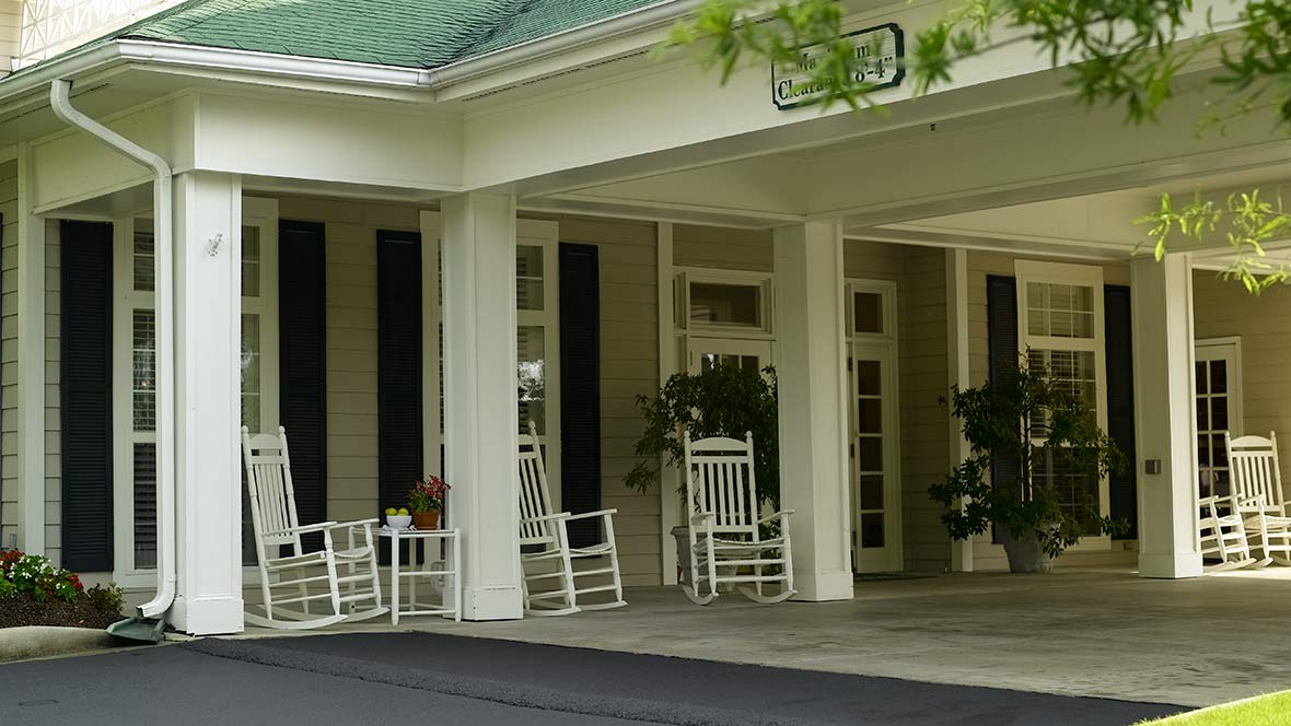 A welcoming entrance with several white rocking chairs on a porch, decorated with plants and supported by columns.