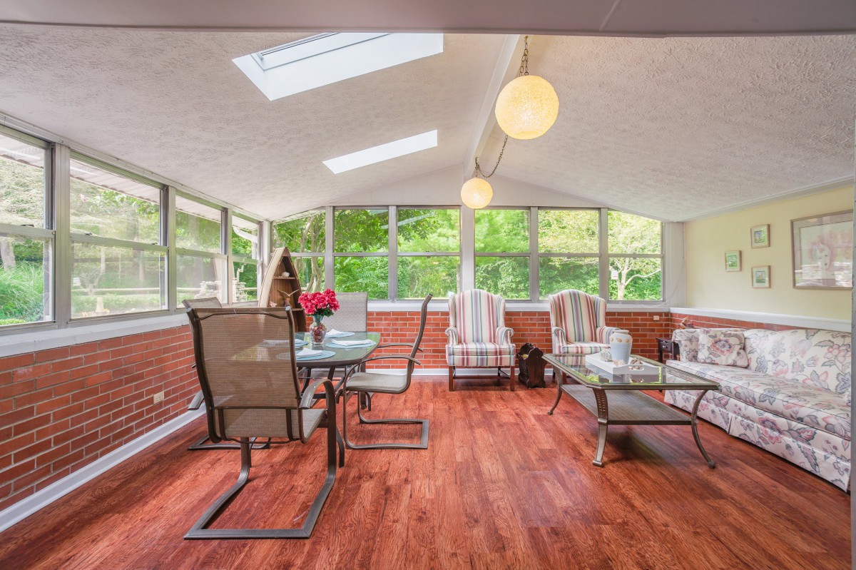 A bright sunroom with skylights, featuring a dining table and comfortable seating arrangements on a wood floor.