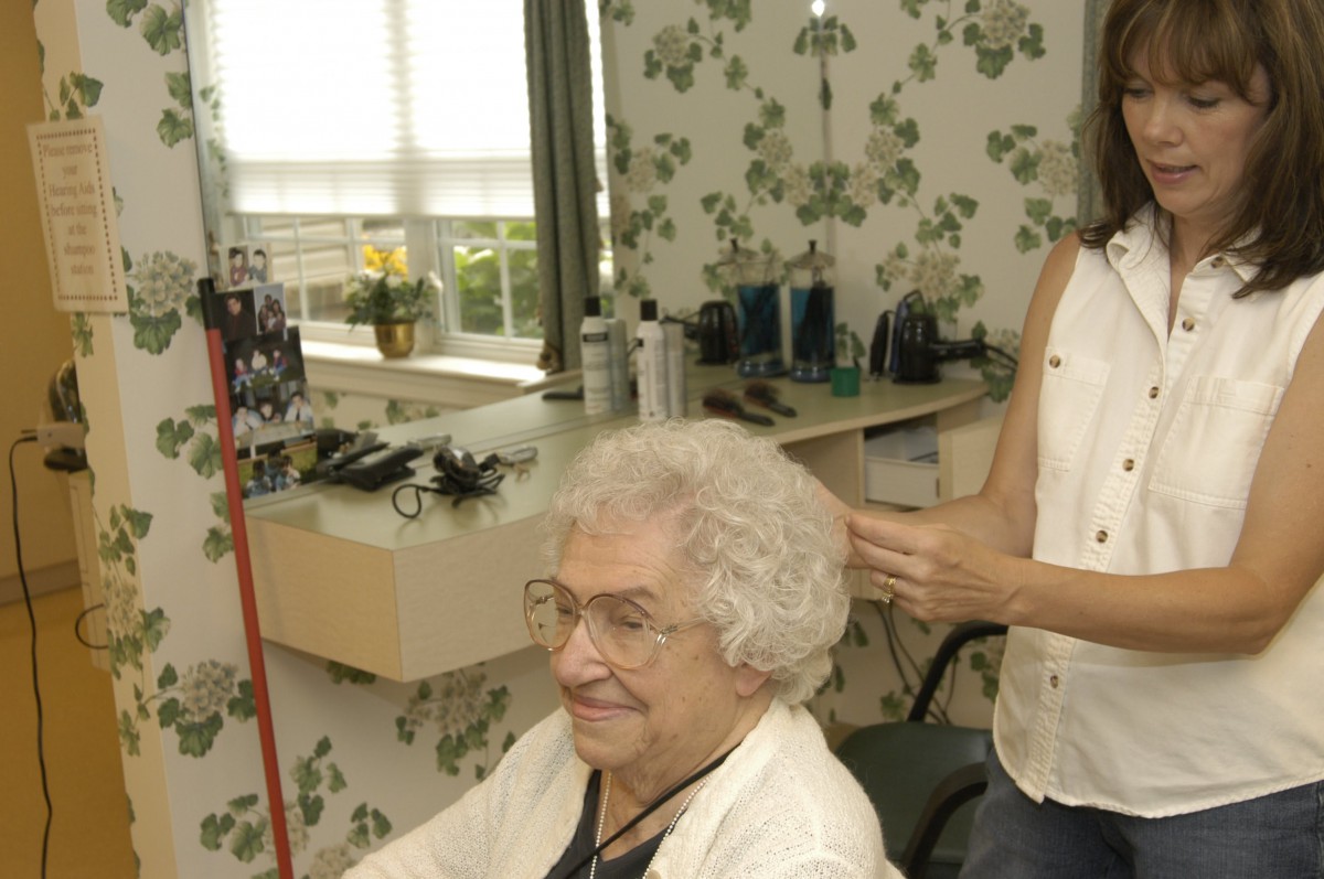 An elderly woman getting her hair styled at a salon, smiling as the stylist works.
