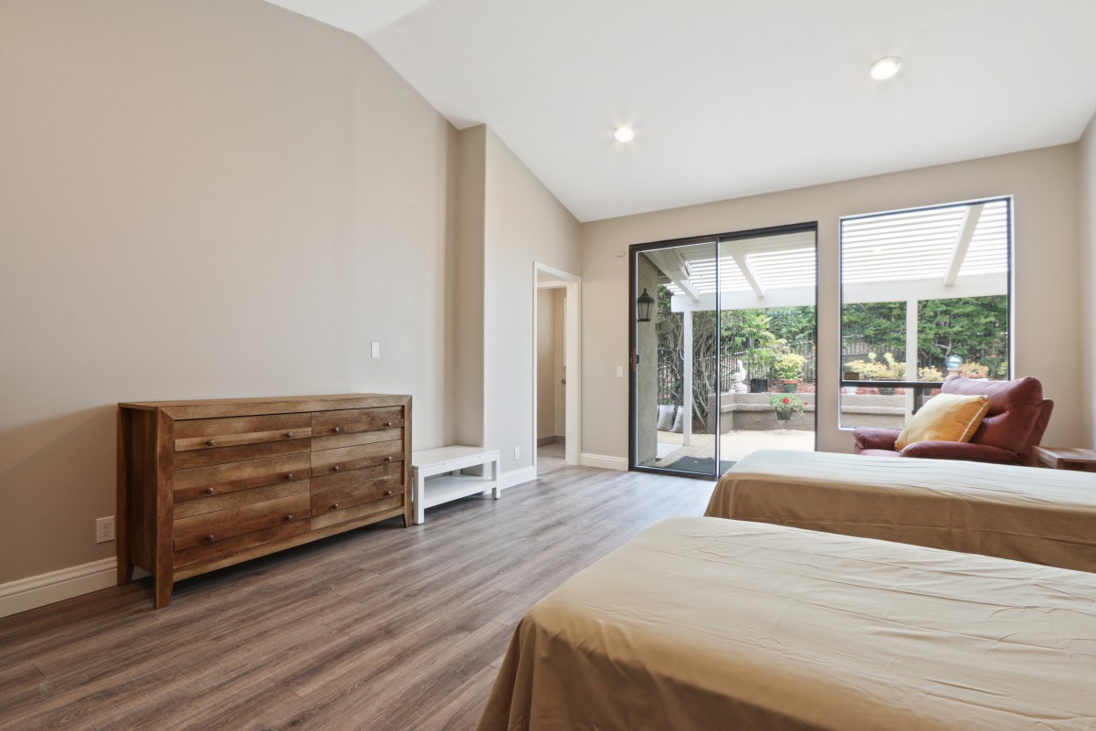 A spacious and minimalist bedroom with wooden flooring, featuring two beds and ample natural light from large windows.