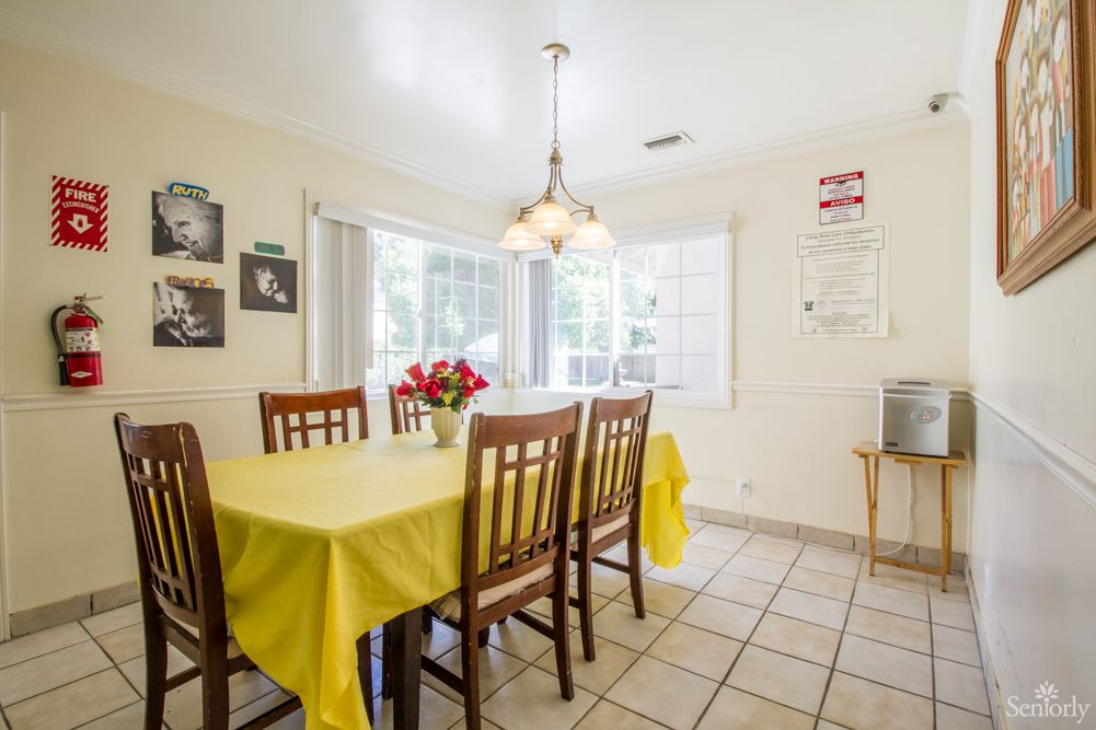 Bright dining room with a yellow tablecloth and wooden chairs, featuring a cozy chandelier and natural light from large windows.