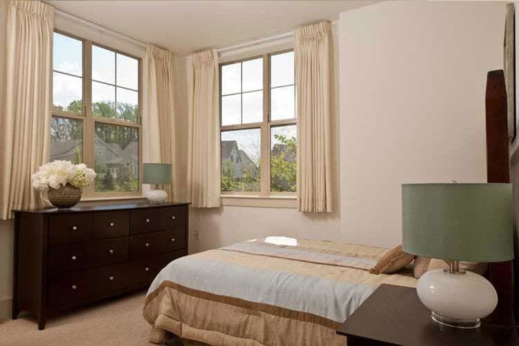 A cozy bedroom with a neatly made bed, large windows, and a dark wooden dresser.