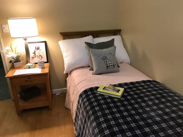 A cozy bedroom with a single bed covered in pink and black bedding, accompanied by a nightstand with a lamp and book.