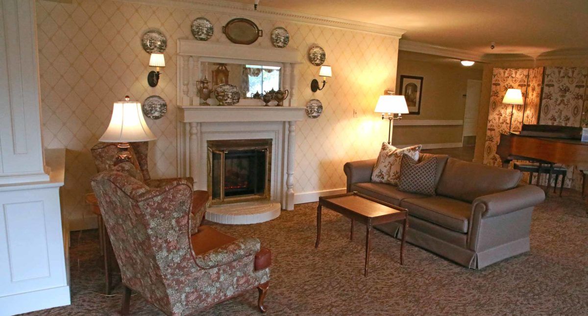 A traditionally styled living room featuring a cozy fireplace, seating area with a sofa and armchair, and decorative elements like wall-mounted plates and warm lighting.