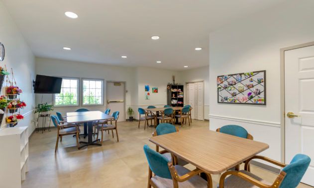 A spacious dining area with several tables and chairs, brightly lit by large windows and overhead lights, featuring artwork on the walls.