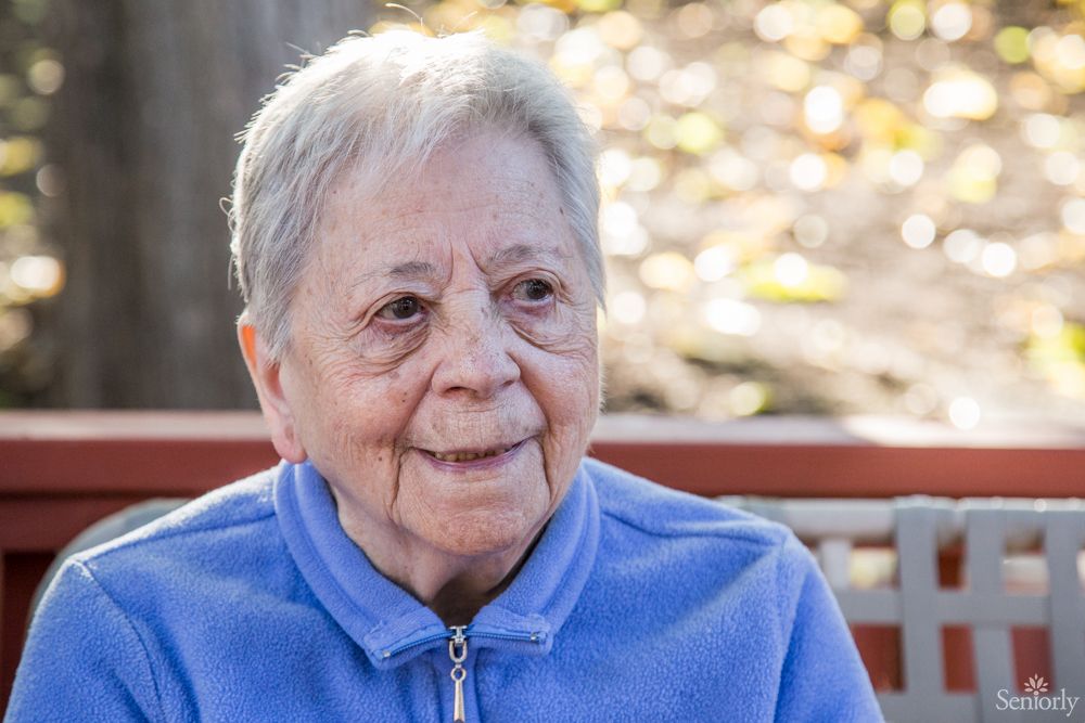 An elderly woman smiles while sitting outdoors in a blue jacket.
