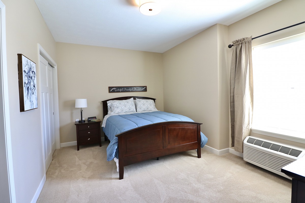 A neatly arranged bedroom with a wooden bed frame, blue bedding, and sheer curtains allowing light to fill the room.