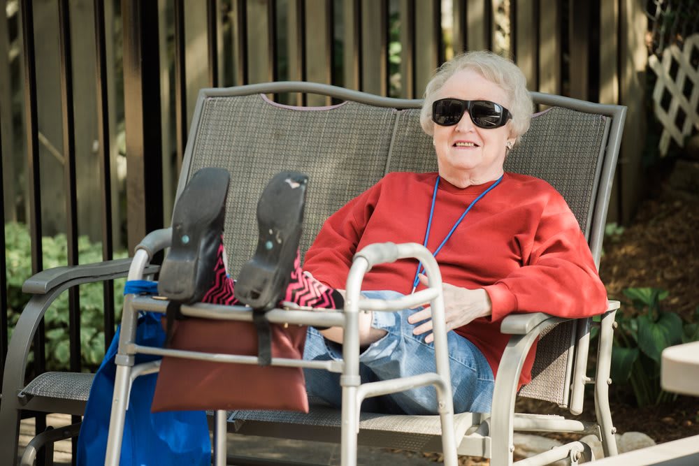 An elderly person wearing sunglasses relaxes outdoors in a chair, with a walker nearby.