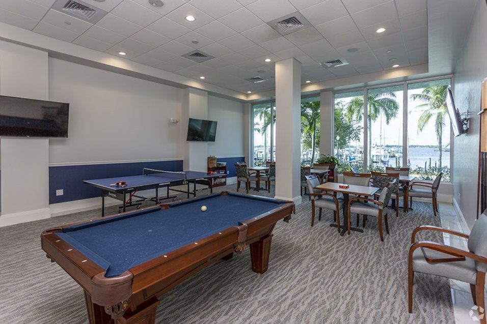A modern game room featuring a pool table, tables, and chairs with natural light streaming through large windows.