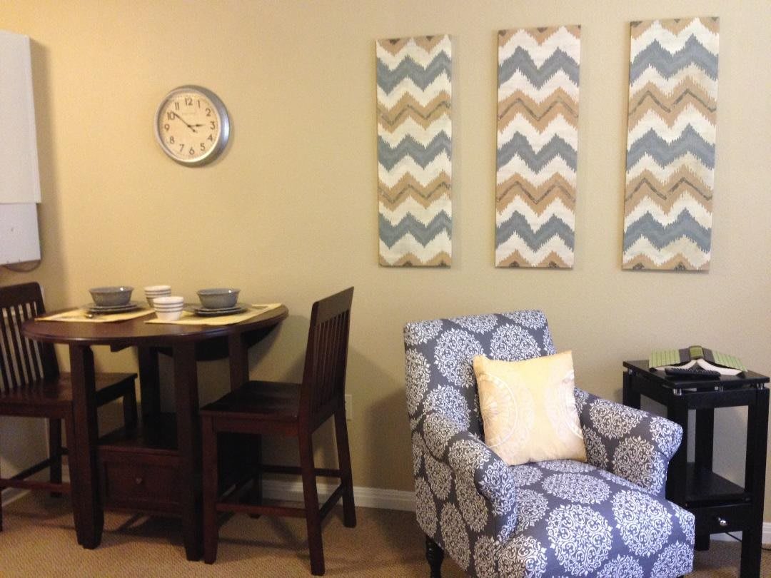 A cozy living room with a patterned armchair, wall art, a clock, and a small dining table set.