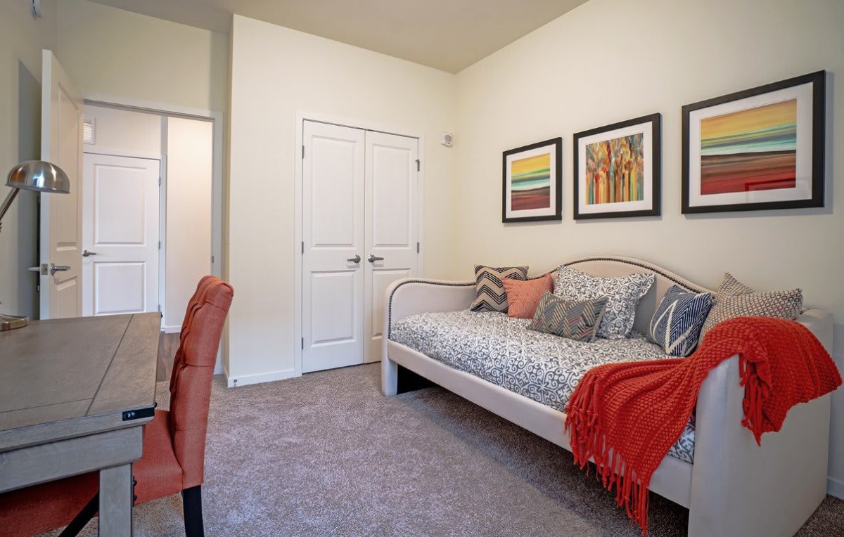 A cozy bedroom featuring a daybed with patterned bedding, accented by colorful pillows and a blanket. The room is decorated with framed artwork on the wall.